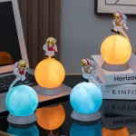 Conjunto de figuras de astronautas com capacetes dourados, cada uma com uma luz de cor diferente, decorando uma mesa com livros e computador ao fundo.