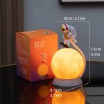 Candeeiro de mesa em forma de lua com astronauta em cima, iluminação amarelada, acompanhado de caixa de embalagem laranja com ilustração do produto.