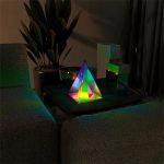 Pirâmide holográfica com luzes RGB coloridas em ambiente escuro, colocada numa mesa junto a plantas e objetos decorativos.