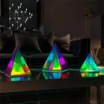 Três pirâmides holográficas de cores vibrantes em diferentes tamanhos, refletindo luzes coloridas numa superfície escura, com fundo de sala moderna.