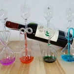 Conjunto de quatro garrafas de vidro com líquidos coloridos, cada uma com tubo de vidro ligado, e uma garrafa de vinho ao fundo.