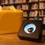 Dispositivo de visualização com ecrã circular mostrando uma imagem de uma figura humana, colocado numa mesa de madeira com uma grande caixa amarela ao lado, fundo de estantes de livros.