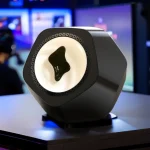 Altifalante com forma hexagonal, superfície preta brilhante e alto-falante circular com detalhe preto no centro, em ambiente de loja de gadgets.