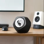 Altifalante portátil preto com logótipo iluminado, colocado numa secretária de madeira junto a um monitor e colunas de som.