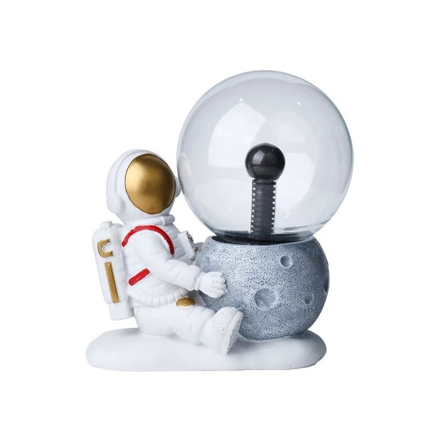 Estátua de astronauta sentado ao lado de uma lâmpada de plasma em forma de planeta, com detalhes dourados no capacete e roupas brancas.