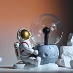 Miniatura de astronauta de brinquedo sentado ao lado de uma lâmpada de plasma, com fundo neutro e iluminação suave.