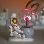 Lâmpada de plasma com efeito de relâmpagos, acompanhada por uma figura de astronauta de brinquedo, em uma superfície branca com fundo desfocado.