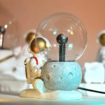 Lâmpada de plasma com esfera de plasma brilhante, base de cimento e figura de astronauta de brinquedo ao lado, fundo desfocado com outros brinquedos espaciais.