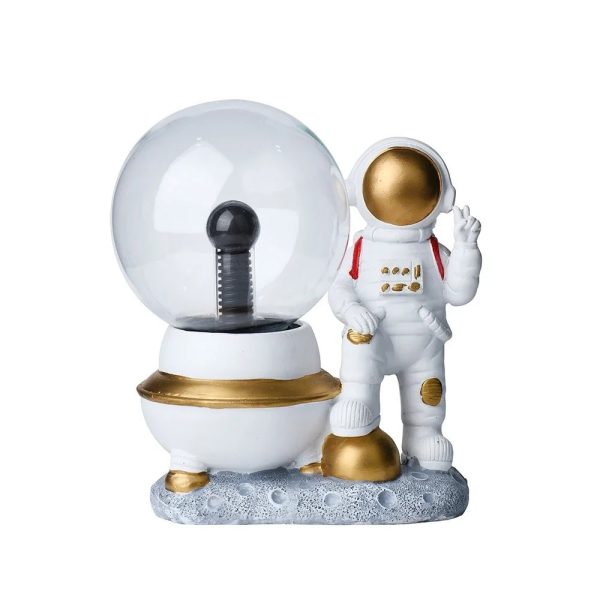 Figura decorativa de astronauta com traje branco, detalhes dourados, junto a um globo de plasma com base branca e dourada, sobre uma superfície de imitação de lua.