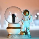 Miniatura de astronauta com capacete dourado ao lado de uma lâmpada de plasma decorativa, ambos em base de rocha, com fundo desfocado de luzes brilhantes.