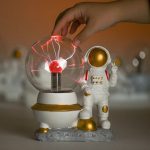Lâmpada de plasma com efeito de relâmpagos, acompanhada de figura de astronauta com capacete dourado, em base decorativa.