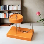 Dispositivo de demonstração com esfera de aço a rolar por um percurso curvo em base laranja, ao lado de uma rosa em vaso.
