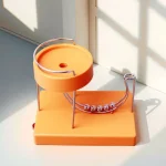 Bola de metal prateada rolando por um percurso curvo de metal, com base de plástico laranja, perto de uma janela com luz natural.