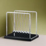 Pêndulo de Newton com bolas de metal suspensas por fios finos, em estrutura de suporte metálico, sobre base preta, contra fundo verde claro.