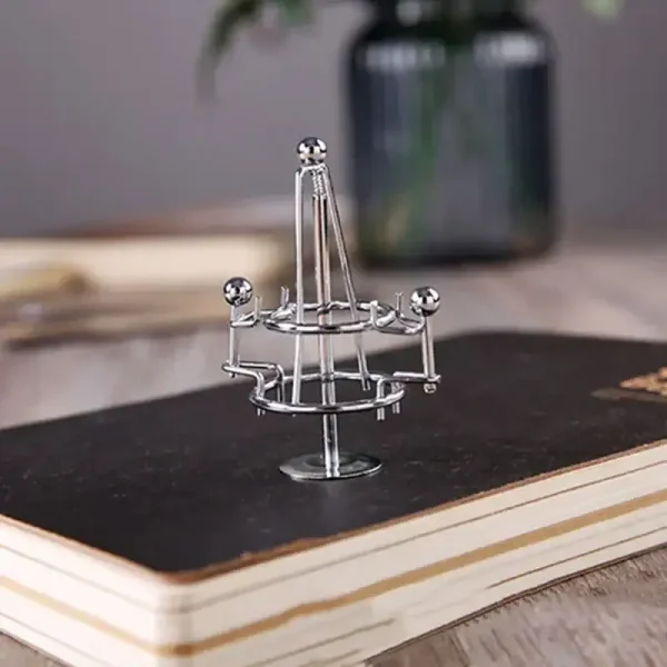 Estrutura de plasma de vidro com esferas metálicas em diferentes alturas, colocada sobre um livro preto em uma mesa de madeira, com uma planta ao fundo.