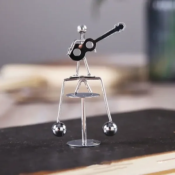 Pequena figura de metal com corpo de esferas, braços e pernas finos, segurando uma guitarra preta, em uma superfície de madeira com fundo desfocado.