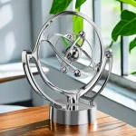 Modelo de átomo em metal prateado com órbitas e partículas, decorativo para ciência ou educação, colocado numa mesa de madeira perto de uma janela com plantas verdes ao fundo.