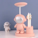 Luminária em forma de personagem astronauta cor de rosa com cabeça espelhada, acompanhada por uma figura menor de astronauta em branco e suporte com canetas decorativas.