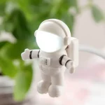 Mini astronauta branco com capacete espelhado, detalhes em preto, pendurado por uma corda, fundo desfocado de plantas verdes.