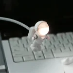 Miniatura de astronauta com luz LED acesa na cabeça, conectado a cabo flexível, sobre teclado de computador.