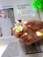Prisma de cristal transparente refletindo luz colorida, segurado por uma mão sobre uma revista de moda e ciência, com destaque para o efeito de dispersão de luz.