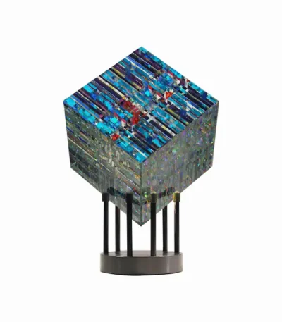 Escultura de vidro com padrões coloridos e textura brilhante, apoiada em suporte de metal com base circular.
