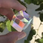 Prisma de cristal transparente com efeito de arco-íris, segurado entre dedos, com folhas verdes ao fundo.