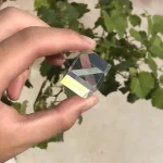 Mão a segurar um prisma de vidro com várias faces, refletindo luz e cores, com plantas verdes ao fundo.