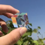 Cubo de cristal transparente com efeito de prisma, refletindo cores do ambiente, segurado por uma mão contra céu azul claro.