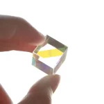 Pequeno cubo de cristal transparente, segurado entre dedos, com efeito de prisma que reflete luz colorida.
