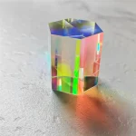 Pequeno cristal prismático com efeito holográfico, refletindo cores vibrantes sob luz direta, colocado sobre uma superfície cinzenta.