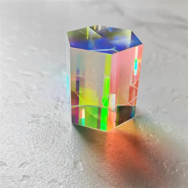 Pequeno cristal prismático com efeito holográfico, refletindo cores vibrantes sob luz direta, colocado sobre uma superfície cinzenta.