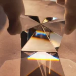 Prisma de cristal com várias faces refletindo luz, criando efeitos de dispersão de cores e sombras no plano de apoio.
