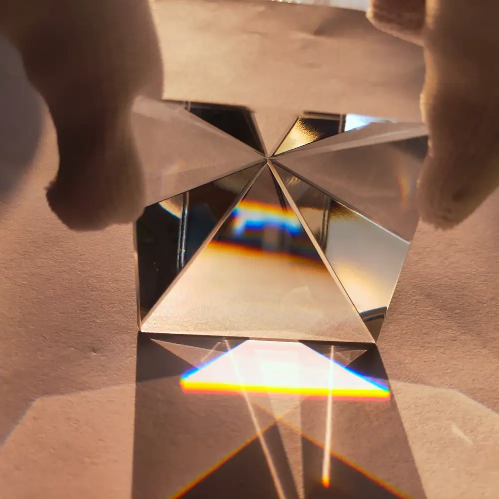 Prisma de cristal com várias faces refletindo luz, criando efeitos de dispersão de cores e sombras no plano de apoio.