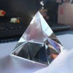 Pirâmide de cristal transparente com múltiplas faces refletoras, colocada sobre uma superfície branca com fundo de escritório.