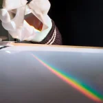 Mão com luva segura um prisma de vidro, projetando um arco-íris de luz sobre uma superfície branca escura.