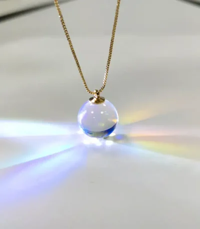 Colar com pingente de bola de vidro transparente que reflete luzes coloridas, suspenso numa corrente dourada fina.