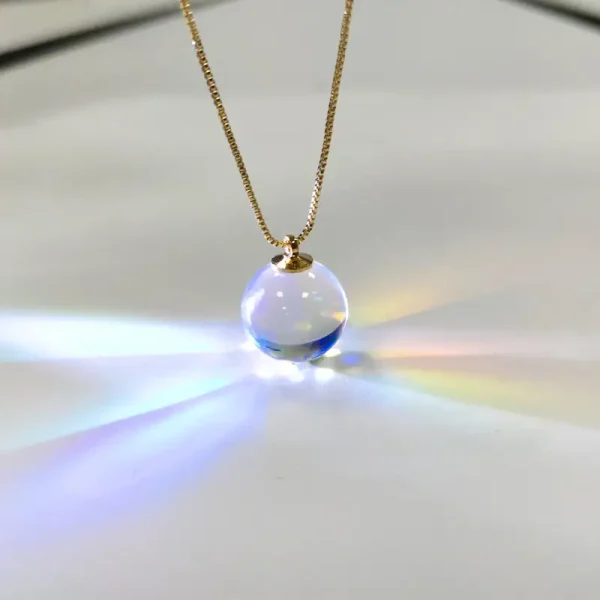 Colar com pingente de bola de vidro transparente que reflete luzes coloridas, suspenso numa corrente dourada fina.