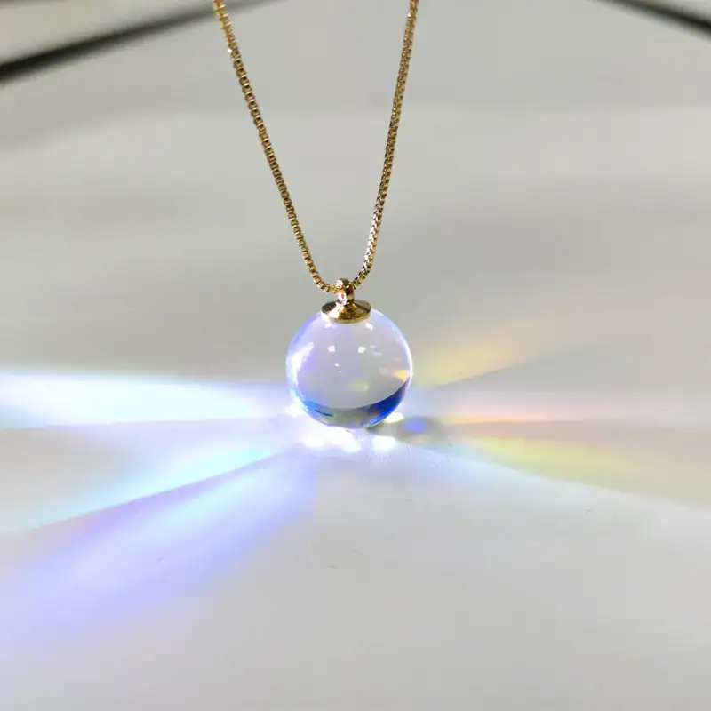Colar com pingente de bola de vidro transparente que reflete luzes coloridas, suspenso numa corrente dourada fina.