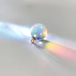 Cápsula de cristal com efeito de luz que projeta cores do arco-íris na superfície, com sombra suave ao lado.