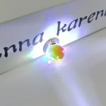 Lâmpada de plasma com efeito de luz colorida, colocada ao lado de uma placa com nome personalizado em letras pretas.