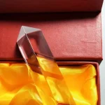 Lâmpada de plasma transparente com base metálica, exibida numa caixa com interior de tecido amarelo.