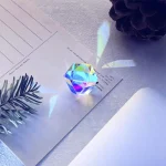 Prisma de cristal com efeito de dispersão de luz, refletindo cores no papel branco ao lado, com uma planta decorativa e uma pedra decorativa ao fundo.