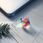Pequeno prisma de cristal com forma de cubo, refletindo luz e criando um arco-íris de cores na superfície de papel branco ao lado de uma folha de papel e uma folha de pinheiro.