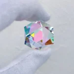 Pequeno cristal de prisma com múltiplas faces refletindo luz, criando efeitos de cores do arco-íris, segurado por uma mão com luva branca.