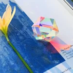 Cubo de vidro transparente com efeito de prisma, refletindo cores do arco-íris, colocado sobre uma superfície azul com uma flor amarela ao lado.