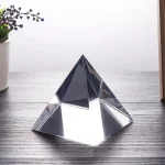 Pirâmide de cristal transparente com várias faces, colocada numa superfície de madeira clara, com uma planta ao fundo à esquerda.