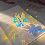Estrela holográfica com cores vibrantes refletindo luz sobre página de livro com texto em francês.