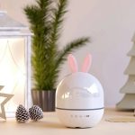 Projetor de galáxia com forma de ovo e orelhas de coelho, em ambiente decorativo com plantas e objetos de decoração.