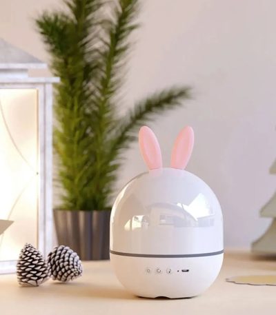 Projetor de galáxia com forma de ovo e orelhas de coelho, em ambiente decorativo com plantas e objetos de decoração.